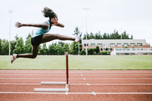 A female athlete in sportswear is mid-air, jumping over a hurdle on a track, with green grass and a building in the background.
