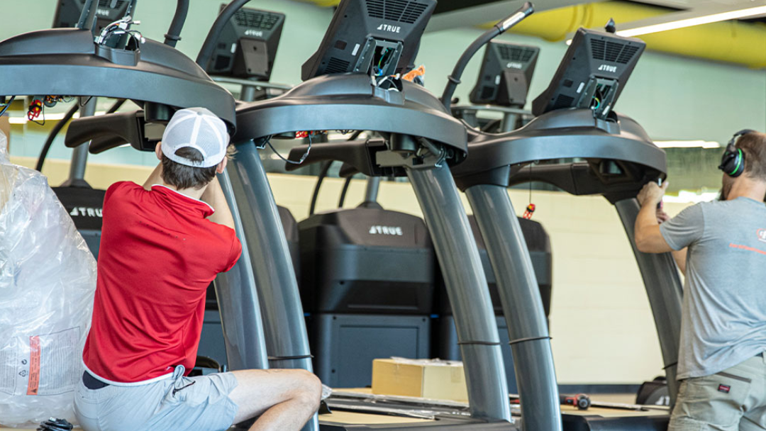 Two men are installing or repairing treadmills in a gym. One man in a red shirt and cap kneels beside a treadmill, while the other, wearing headphones, works on a different treadmill. Various tools and boxes are nearby.