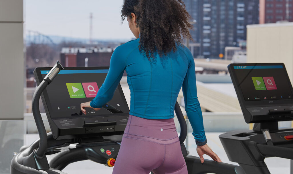 A woman in a blue long-sleeve top and purple leggings uses the touchscreen display of a treadmill on an indoor balcony, with buildings and a bridge visible through the windows behind her.