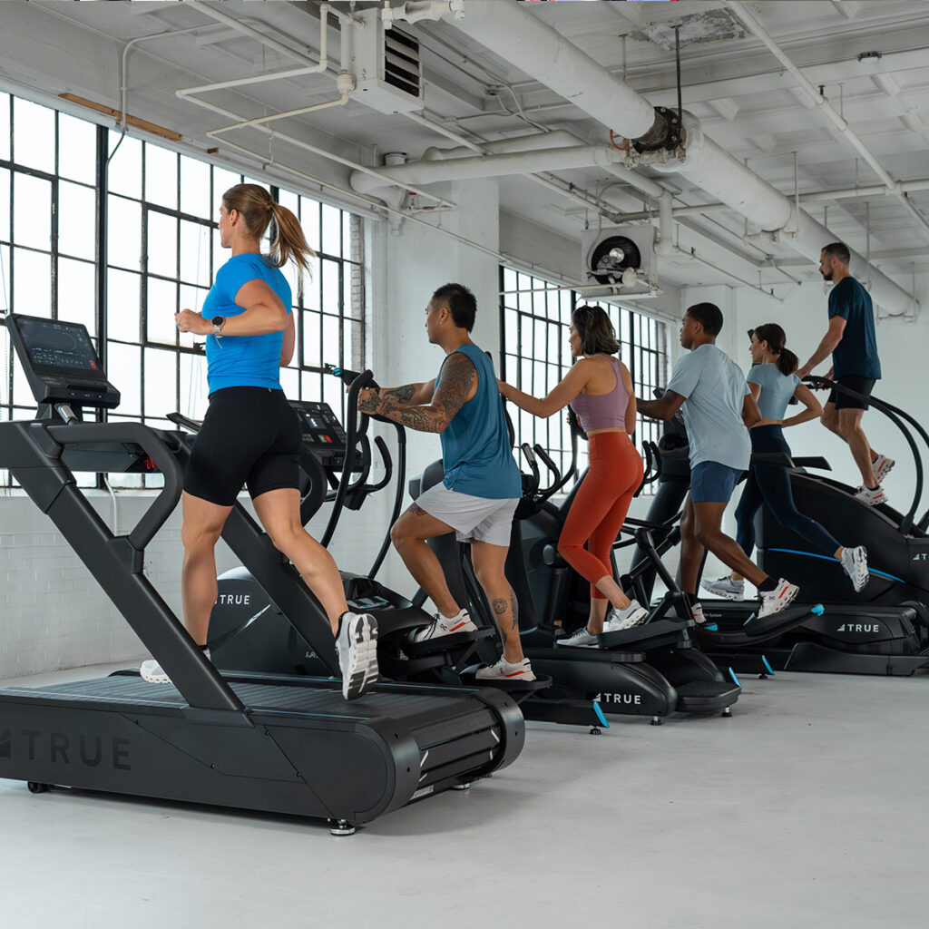 A group of six people exercise indoors on treadmills and stair climbers by large windows, wearing athletic clothing in a brightly lit gym.