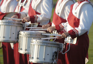 Four marching band members in red and white uniforms play snare drums outdoors, holding drumsticks poised above the drumheads, with only their torsos and arms visible.