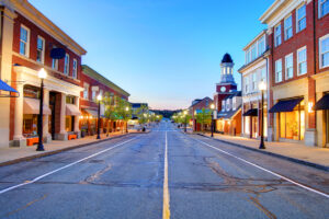 A quiet, empty main street lined with brick buildings, shops, and street lamps at dusk. The sky is clear and blue, and a clock tower is visible in the distance.