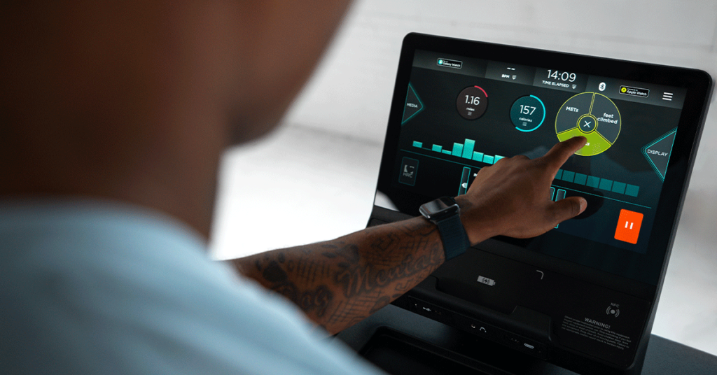 A person wearing a smartwatch interacts with a touchscreen fitness device, selecting workout options displayed on a digital interface showing stats like heart rate and exercise duration.