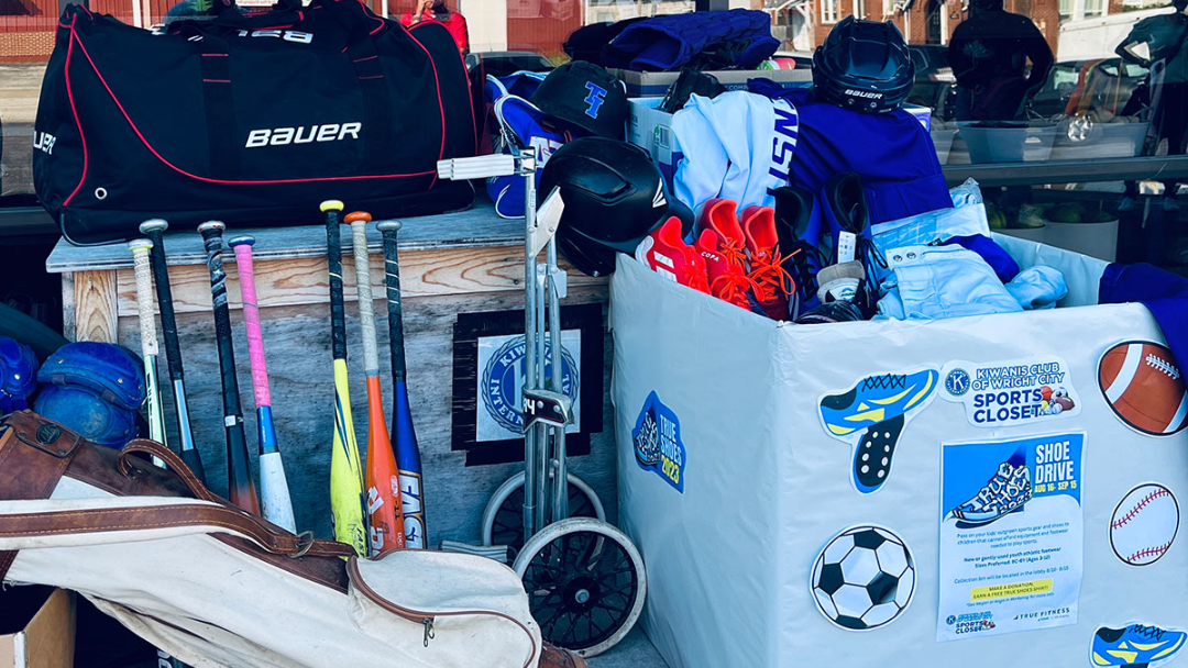 A collection bin filled with sports equipment and shoes sits beside bags, bats, balls, gloves, and helmets, showing items donated to a sports gear drive. A sign on the bin promotes the donation event.