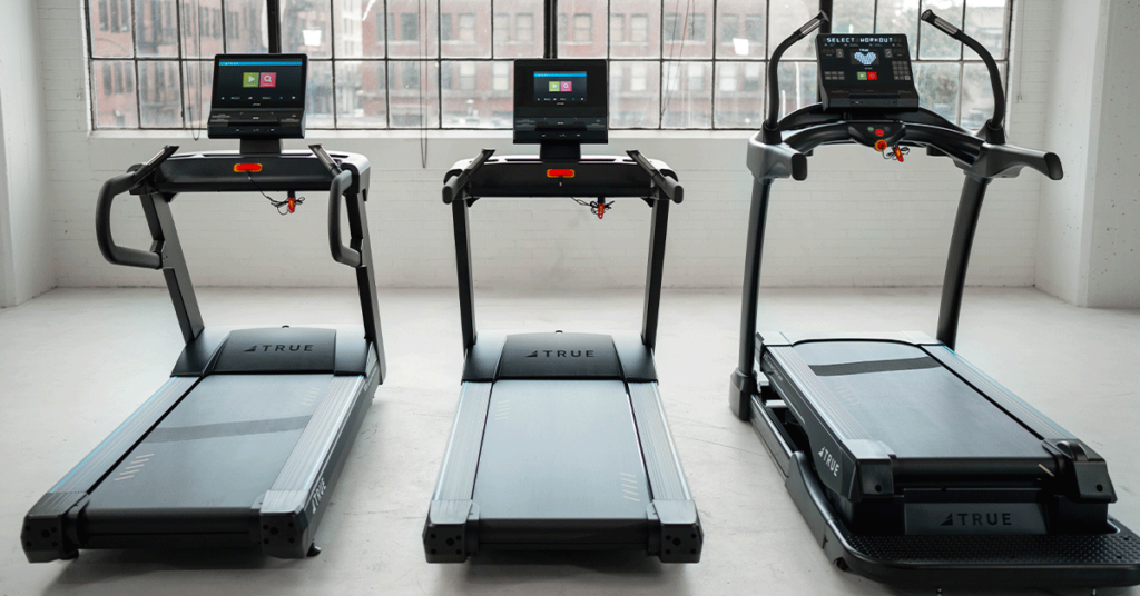 Three modern treadmills with digital screens are positioned side by side in a bright room with large windows and a city view. The treadmills are black and display the brand name TRUE on the base.