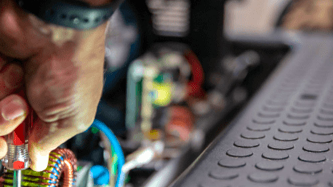 A close-up of a persons hand using a screwdriver to work on electrical components, with colorful wires and circuitry visible, next to a textured, rubber-like surface.