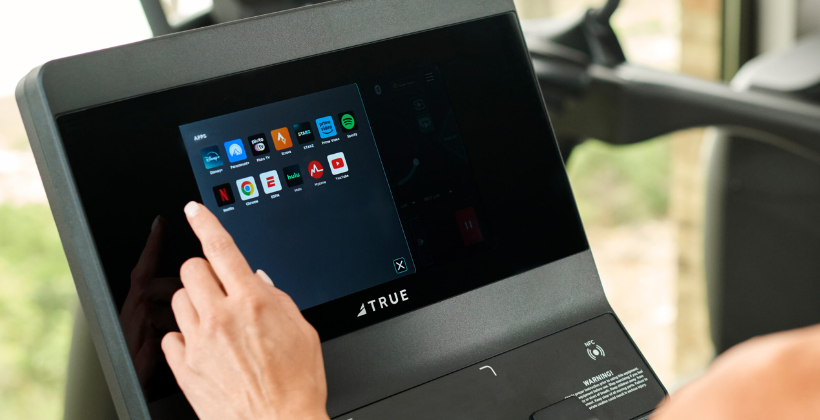 A person’s hand is touching the screen of a 66体育 - TRUE Fitness让运动更简单_官方网站 66体育 66体育 machine, selecting from various apps like Netflix, YouTube, Spotify, and others displayed on the touchscreen interface.