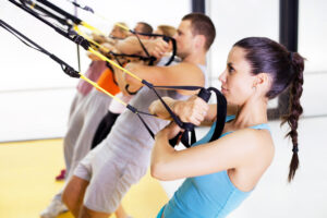 A group of people exercise in a gym, using suspension straps attached above them. They pull the straps while leaning back, engaging their arms and core. The focus is on a woman in front, wearing a light blue tank top.