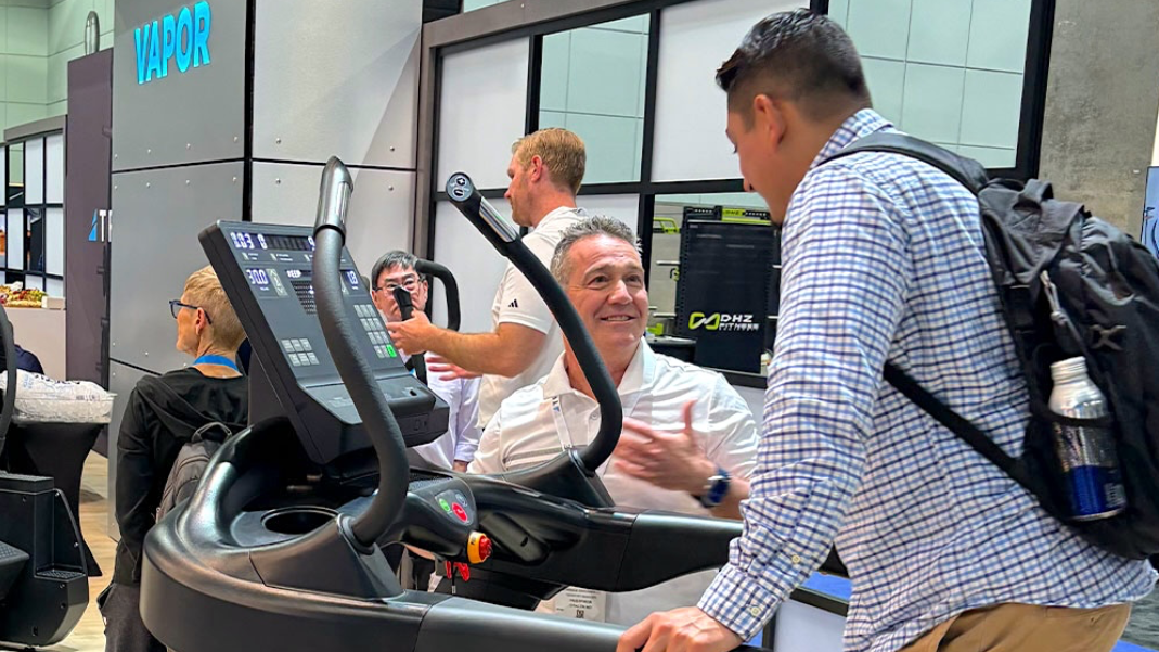Two men talk enthusiastically near a treadmill at a trade show booth. One is seated on the treadmill, gesturing, while the other stands with a backpack. Others interact in the background near displays and signage.