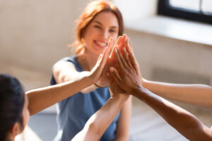 A group of people sitting in a circle smile and stack their hands together for a team high five, showing unity and support.