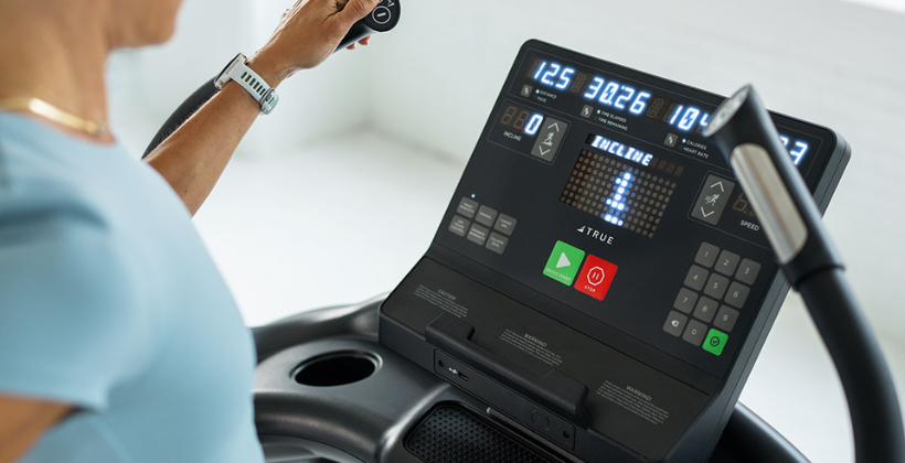 A person uses a treadmill, adjusting the controls on the digital display panel showing workout data such as speed, time, distance, and incline. The person is wearing a light blue shirt and a watch.