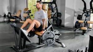 A man wearing a gray T-shirt and black shorts is using a leg press machine in a gym. The gym has various equipment and large windows with trees visible outside.